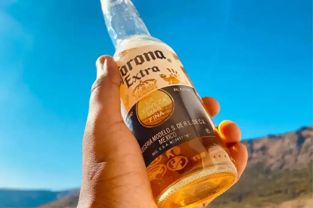 Mão segurando garrafa gelada de Corona Extra com paisagem ensolarada ao fundo