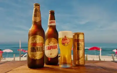 Garrafas, lata e copo de Itaipava 100% Malte e Pilsen em mesa de madeira na praia