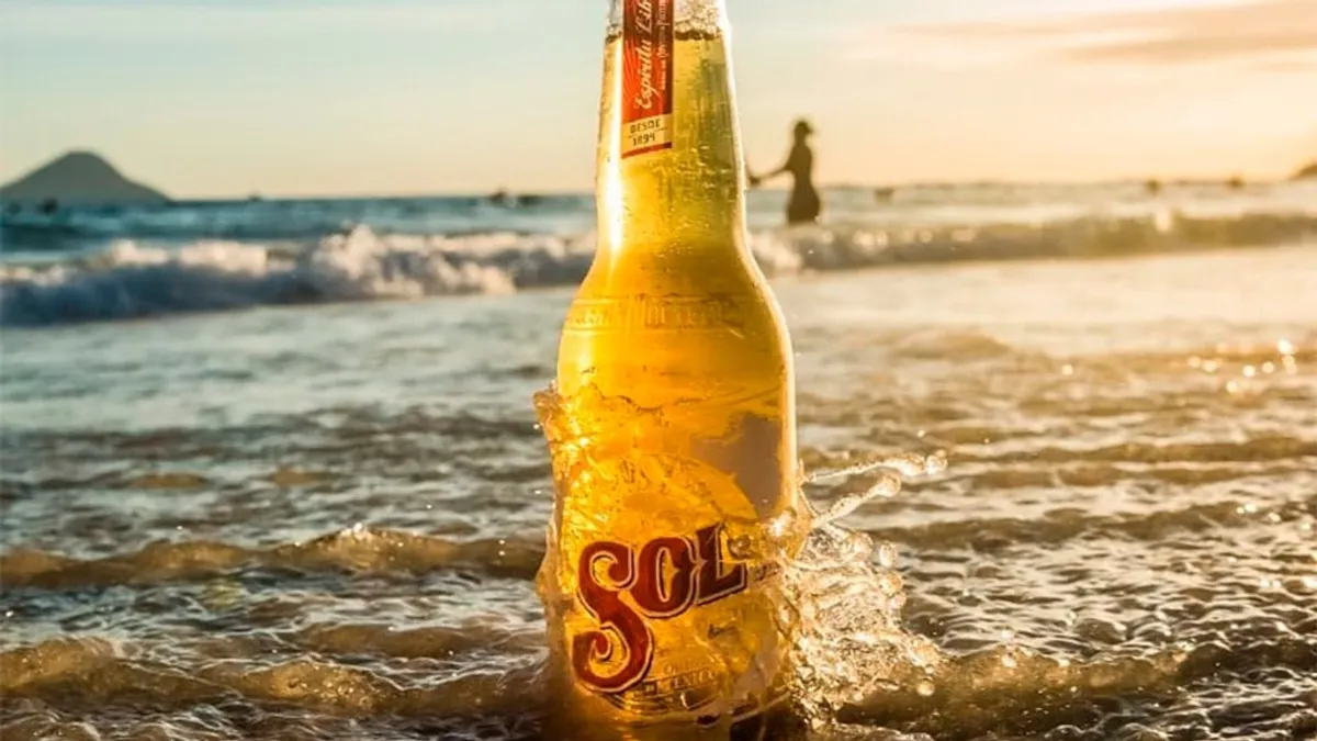 Garrafa transparente de cerveja Sol na areia com onda ao pôr do sol