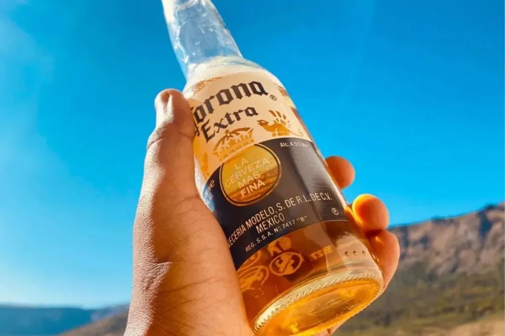 Mão segurando garrafa gelada de Corona Extra com paisagem ensolarada ao fundo